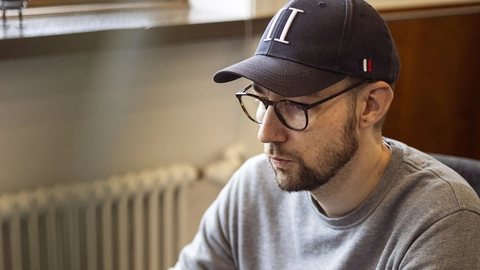 En mand sidder ved et skrivebord og arbejder på en bærbar computer, iført en grå sweater og en baseballkasket, med en mobiltelefon og høretelefoner på bordet.